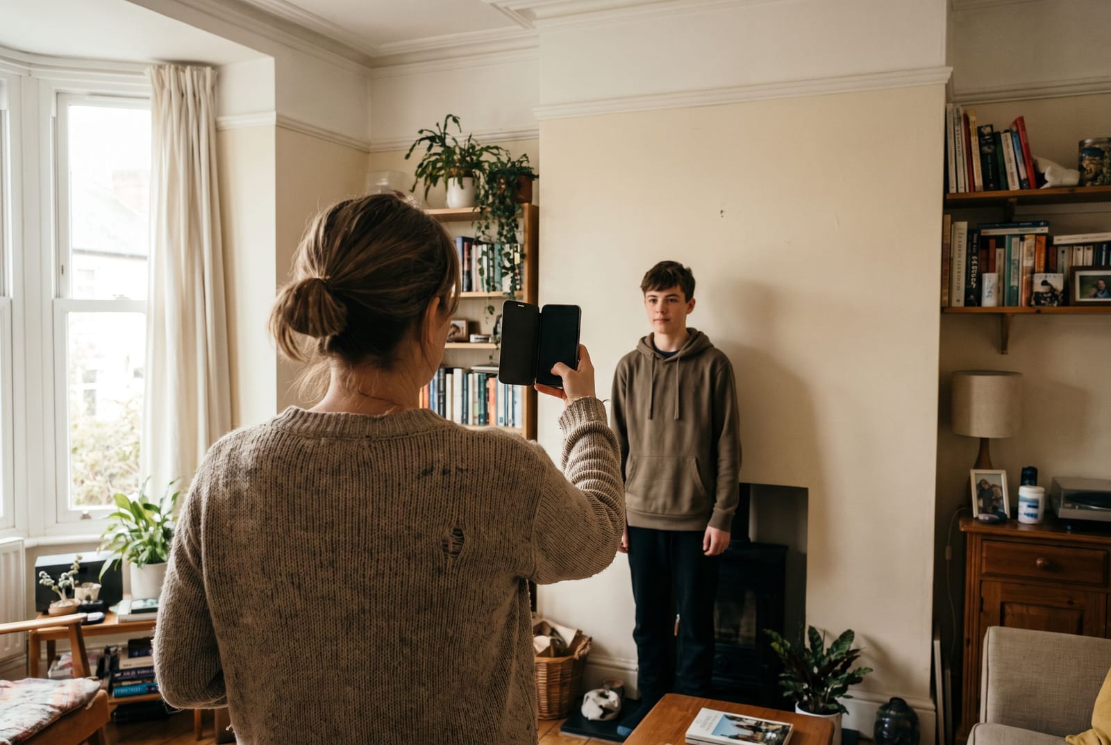 A woman photographing her teenage son against a plain wall at home, using a phone
